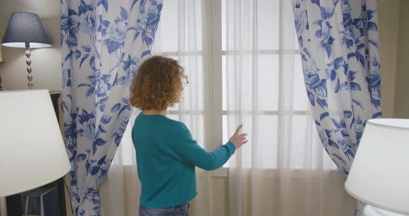 Young Woman Opening Curtains and Looking Out Window at Home.