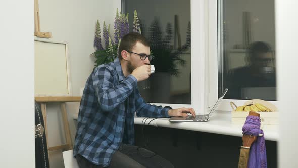 Young business man using laptop computer at remote home workplace on balcony and drinking coffee