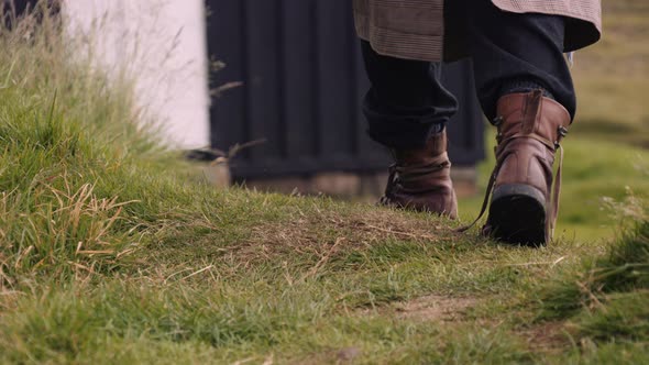 Mans Feet in Leather Boots Walks on Grass Towards Little Black House