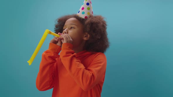 Cheerful African American girl in birthday party hat blowing yellow pipe