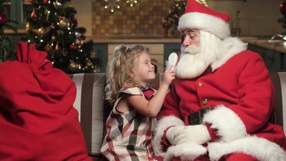 Little Girl on Christmas Night Is Sitting and Combing Santa Claus White Beard