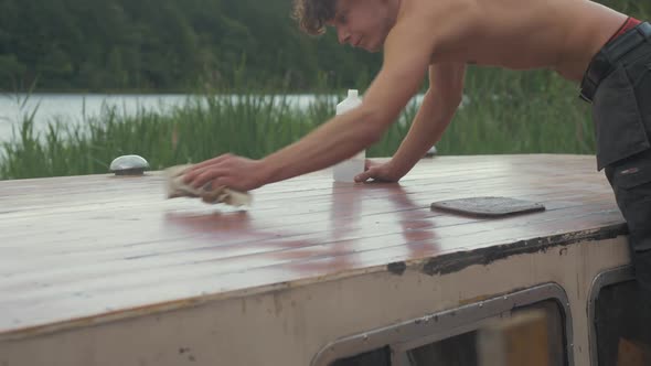 Young carpenter brushing down sanded roof planking of old wood boat using acetone on cloth in hot Su