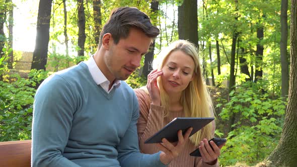 A Young Attractive Couple Works on a Smartphone and a Tablet on a Bench in a Park on a Sunny Day