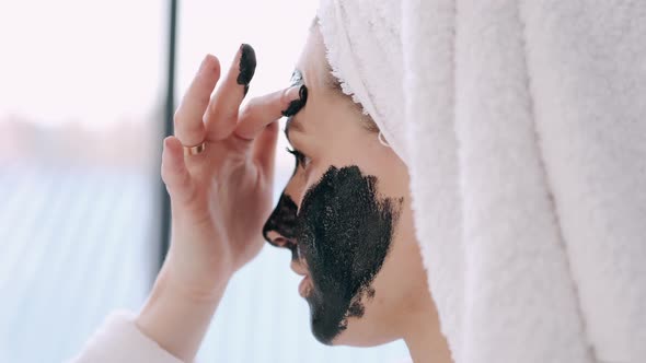 Pretty Woman with Wrapped Hair in a Towel Is Applying Black Mask on Face