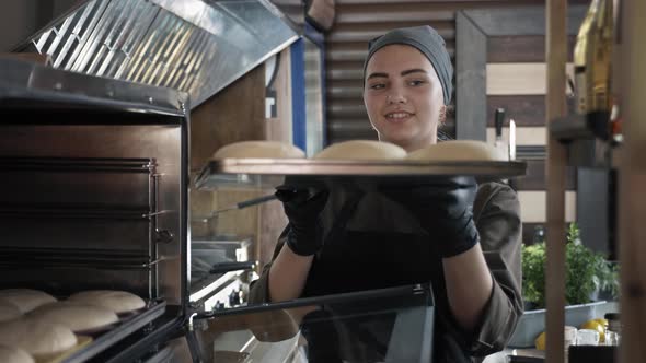 Local Producers, Female Baker in Uniform Puts Baking Sheet with Slices of Dough in Oven To Get Fresh