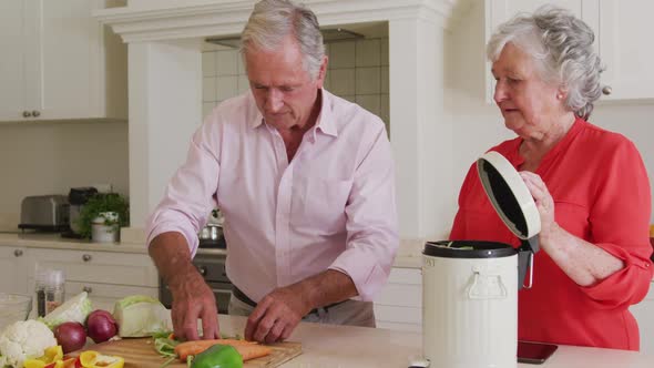 Happy caucasian senior couple in kitchen preparing meal, composting vegetable scraps