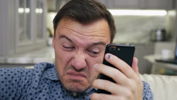 Peering Man in Shirt Looking Into Phone Screen While Sitting at Sofa in Living Room