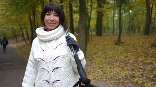 Smiling Caucasian Mature Woman Looking at Camera Standing in Autumn Park with Ski Poles