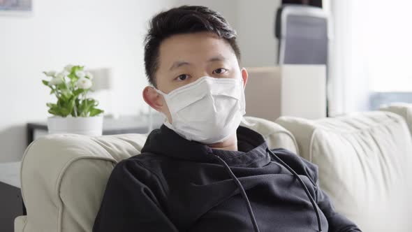 A Young Asian Man in a Face Mask Shows a Thumb Down and Shakes His Head As He Sits on a Couch