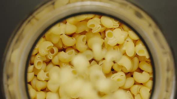 TOP VIEW: Uncooked Curly Macaroni Fall Into Glass Jar On Black Background - Slow Motion