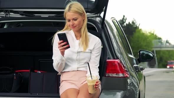 Young Attractive Blond Woman Sits in the Trunk, Works on the Smartphone and Drinks Ice Coffee