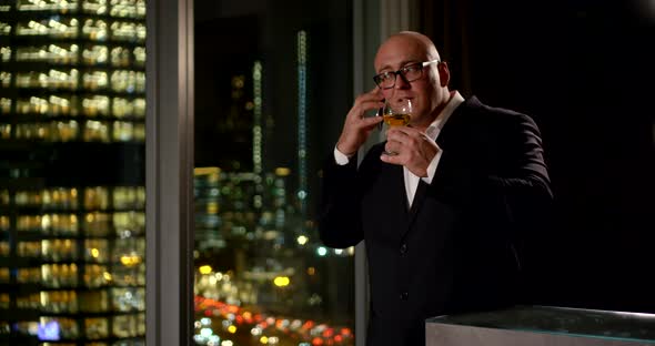 Middle-aged Man in Glasses and a Suit Is Talking on the Phone and Drinking Wine at a Large Window