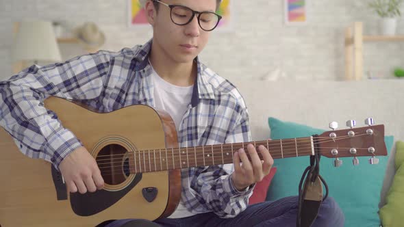 Positive Young Asian Man in Hat and Stylish Glasses with Guitar Looking at Camera Sitting on Sofa in