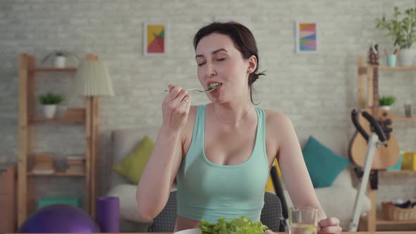 Happy and Joyful Young Sporty Woman Eating Vegetables Sitting at the Table