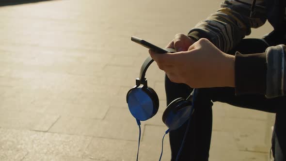 Hands of a Teenage Boy with Smartphone and Headphones  He Chatting with Friends Outdoor