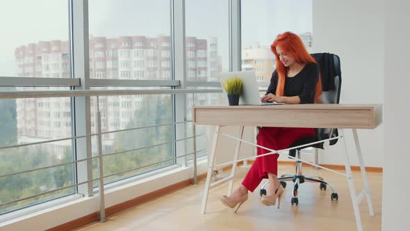 Young Attractive Woman with Red Hair Working in a Bright Office with Panoramic Windows Working on a