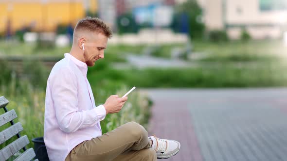 Attractive Hipster Male in Earphones Relaxing Sitting on Bench Listening Music Using Smartphone