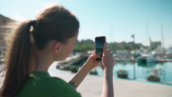 Blonde woman wearing green t-shirt making photo of the seafront on mobile near yachts