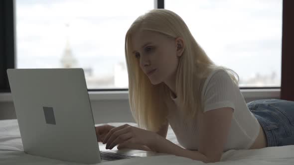 Beautiful Blonde Woman Lies on the Bed with a Laptop