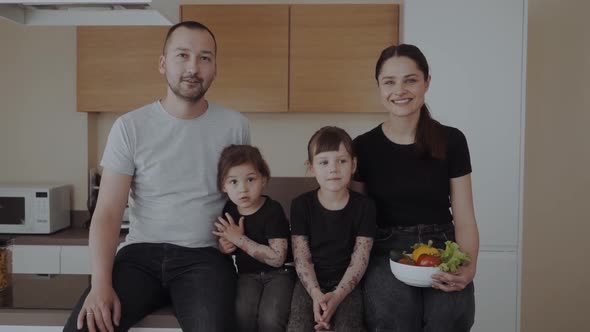 Happy Family in the Kitchen Sitting on the Table with a Bowl of Vegetables and Smiling at the Camera