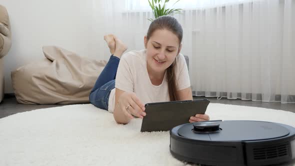 Young Smiling Woman Resting and Browsing Internet on Tablet Computer While Roboto Vacuum Cleaner