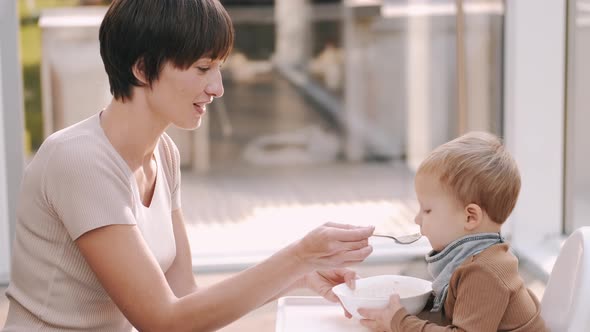 Child Sitting in a Baby Chair and Eating Food with Mother