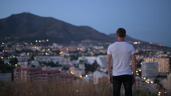 Rear View Of Young Tourist Man With Arms Raised On Top Of The Hill At Night