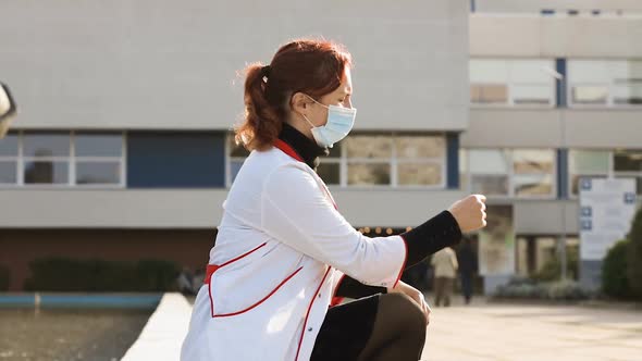 Sad Exhausted Young Doctor or Nurse Sits in Face Mask Near Hospital