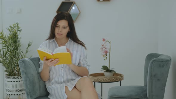 Serious millenial woman in casual clothes reading her planner book and sitting on chair at home