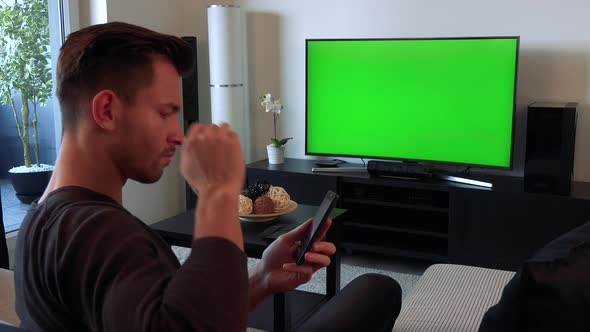 A Man Watches a TV with a Green Screen in a Cozy Living Room and Types on His Smartphone