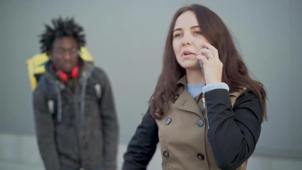 Portrait of Irritated Young Caucasian Woman Talking on the Phone As Blurred African American Courier