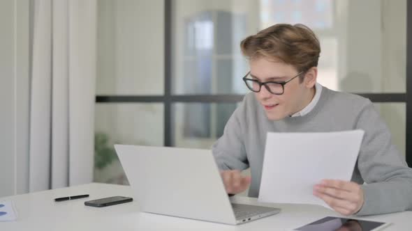 Young Man with Laptop Celebrating Success While Reading Documents in Office