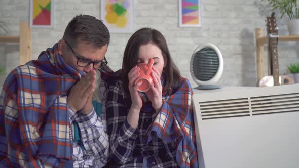 Frozen Young Woman and Man Wrapped in a Blanket in the Living Room Are Heated Next To Electric