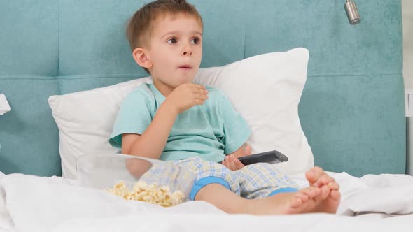 Little Boy in Pajamas Eating Popcorn in Bed at Morning and Switching Cartoons on TV with Remote