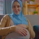 Positive Pregnant Muslim Young Woman in a Headscarf is Sitting on the Sofa and Looking at the Camera - VideoHive Item for Sale