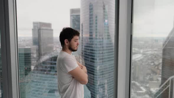 Young Man Thinks and Looks Out the Window in a Modern Apartment Overlooking the City From the Top