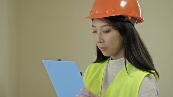 Asian Builder Woman Wearing Protective Clothing and a Helmet is Writing Down Something