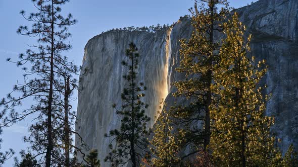 sun sets on yosemite waterfall as mist shines orange in fading sunlight alt