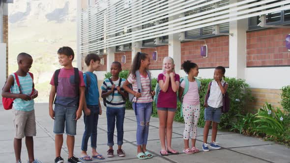 Diverse group of schoolchildren wearing backpacks smiling and standing in a row at school yard