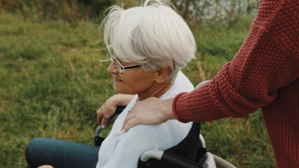 Elderly Mother in Wheelchair with Daughter in Autumn Nature. Walking Along the River