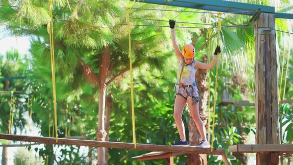 a Cheerful Teen Girl in a Helmet Tshirt and Shorts Climbs in a Rope Park