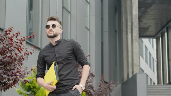 Young Male Businessman Holding a Yellow Folder with Documents and a Smartphone. A Man Reads a