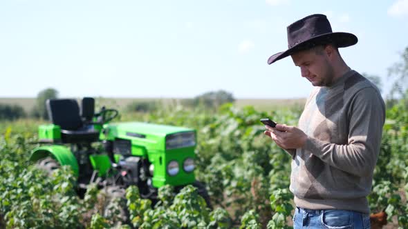 Caucasian Professional Young Farmer Standing with Smartphone on Green Tractor Background