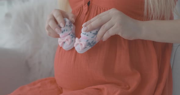 Close-up of Young Caucasian Female Hands Holding Baby Booties and Imitating Stepping on Pregnant