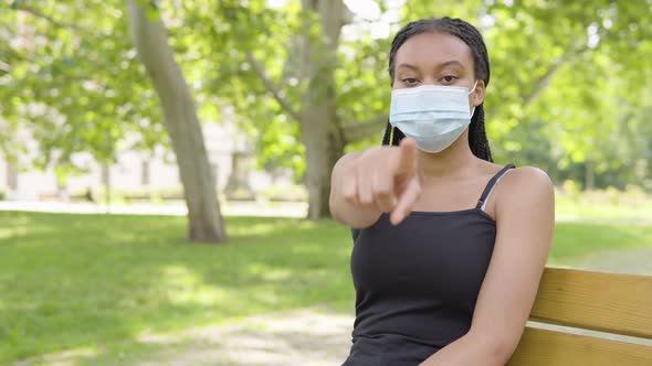 A Young Black Woman in a Face Mask Points at the Camera and Nods As She Sits on a Bench in a Park