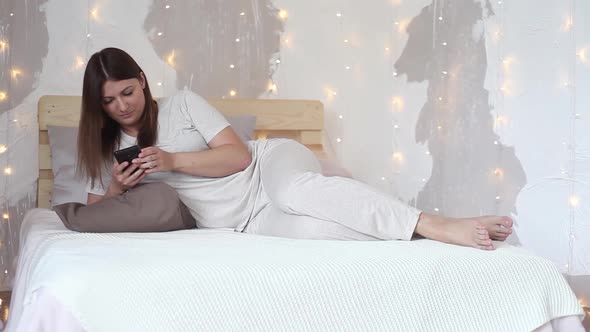 Happy Beautiful Girl in Pajama with Long Hair Enjoys Mobile Phone Lying on Bed at Home