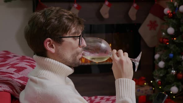 A guy with glasses looking through the phone and drinking some sparkling wine.