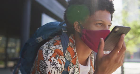 African american man in city, wearing face mask sitting using smartphone in street