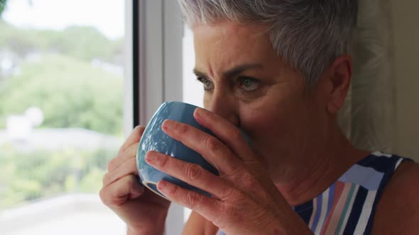 Caucasian senior woman drinking coffee while looking out of the window at home
