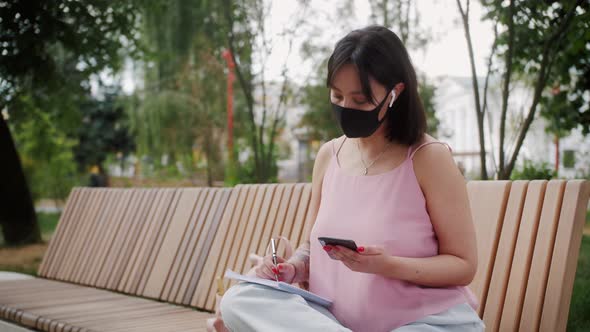 Asian girl wearing protective medical mask in public space at the park using smartphone.